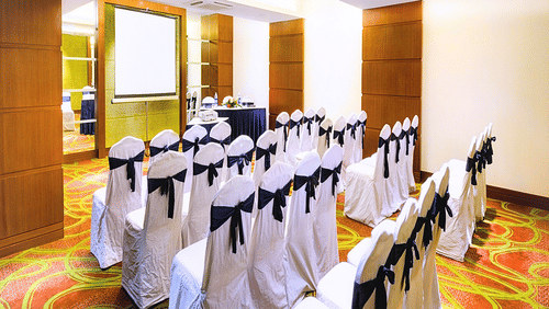 A conference room at Pride Plaza Ahmedabad, featuring rows of chairs with black sashes, a projector screen, and natural light streaming in.